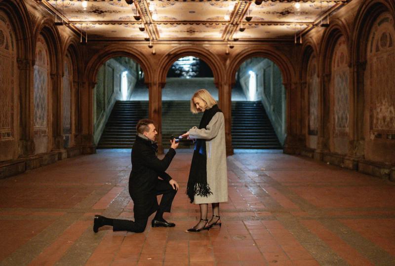 A man is proposing to his partner at Bethesda Terrace in Central Park. He is kneeling with an open ring box in his hand while she smiles and looks down at him. They are standing under the iconic arches with the staircase in the background. The warm lighting and historic architecture create a romantic atmosphere, highlighting this special moment in one of New York’s most famous landmarks.