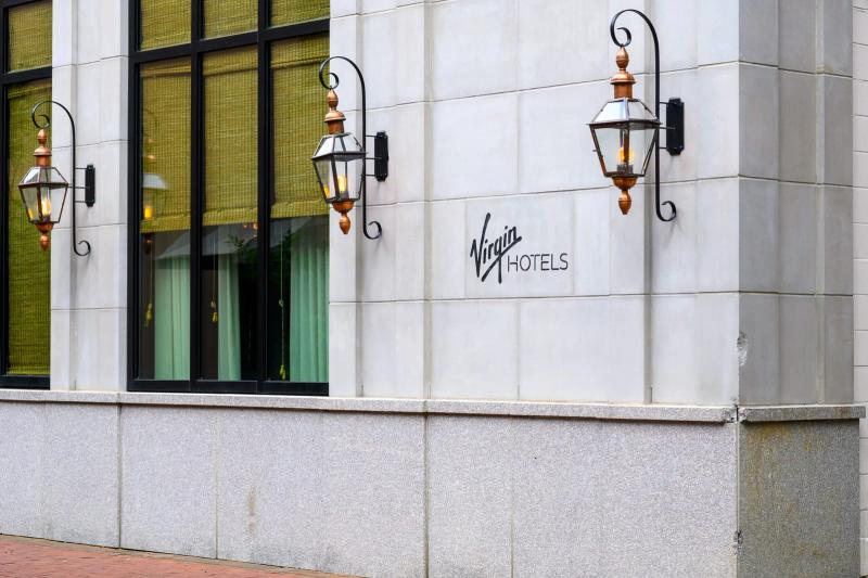 Exterior view of Virgin Hotels New Orleans showing the building’s corner with large windows, decorative wall lanterns, and the Virgin Hotels logo on a light stone facade.