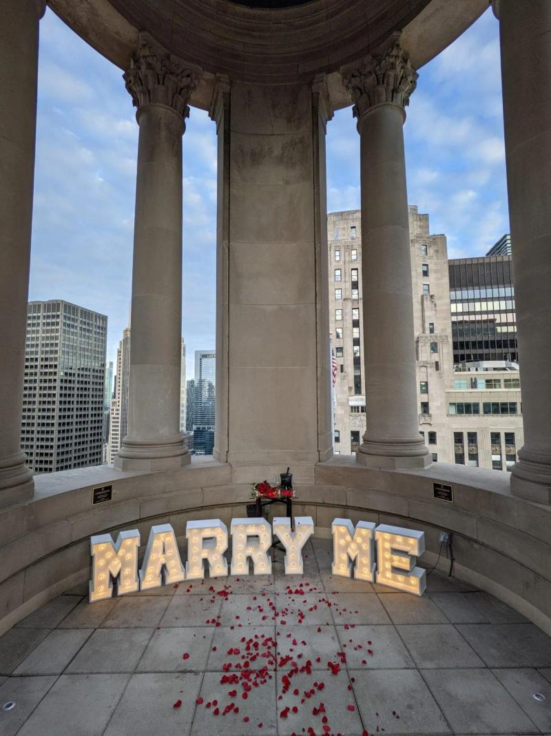 Romantic proposal setup at LondonHouse Cupola in Chicago with illuminated “Marry Me” letters, rose petals, champagne, and skyline view.