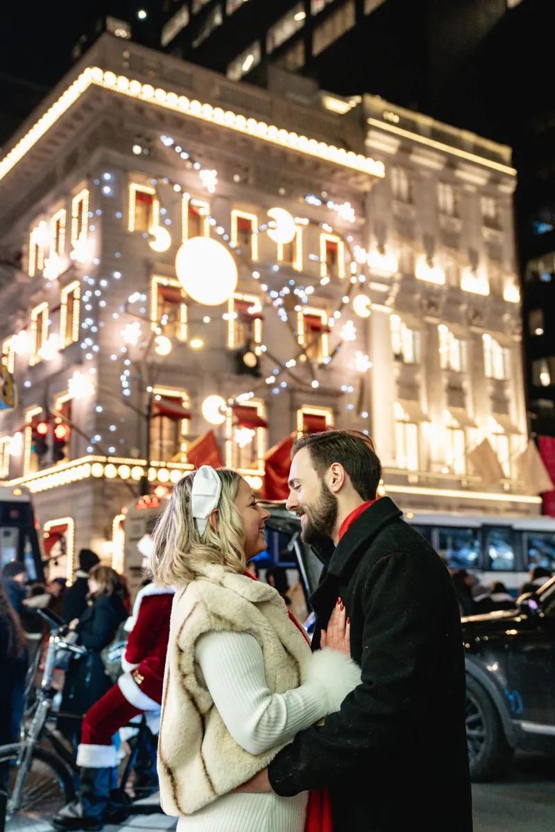 A couple is standing close together on Fifth Avenue in New York during the holiday season. They are smiling at each other while holiday lights, festive store decorations, and busy street activity create a bright and lively backdrop. The woman is wearing a white outfit with a fur vest and a bow headband, and the man is wearing a dark coat with a red scarf. The scene captures a warm romantic moment in the middle of New York’s Christmas atmosphere.