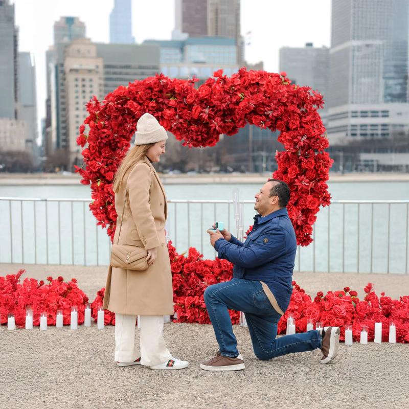 Marriage proposal at Milton Lee Olive Park in Chicago, man on one knee with ring in front of red heart-shaped flower arch by the lake.