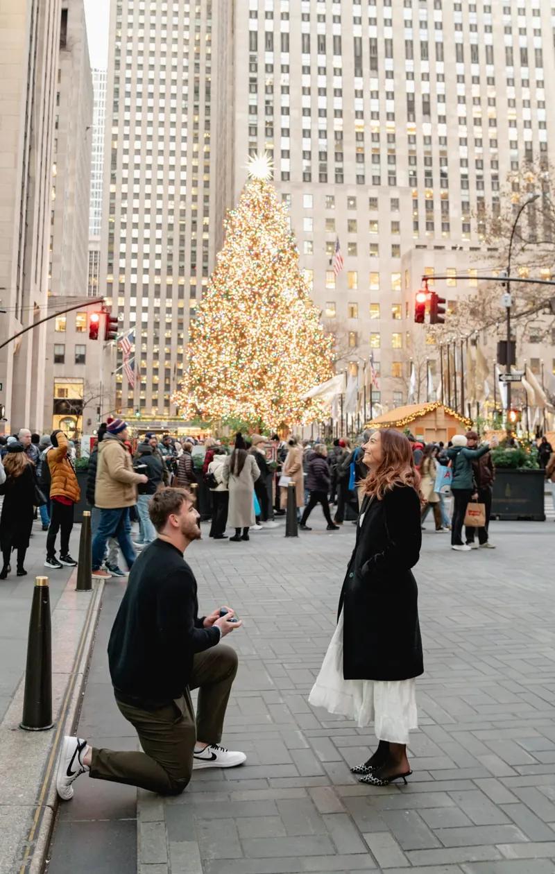 A man proposes to his partner at Rockefeller Center in New York. He is kneeling on the pavement holding a ring box while she smiles at him. In the background the famous Rockefeller Christmas tree glows with lights and crowds of people walk through the festive holiday scene. This moment captures a classic New York City proposal in one of its most iconic winter locations.