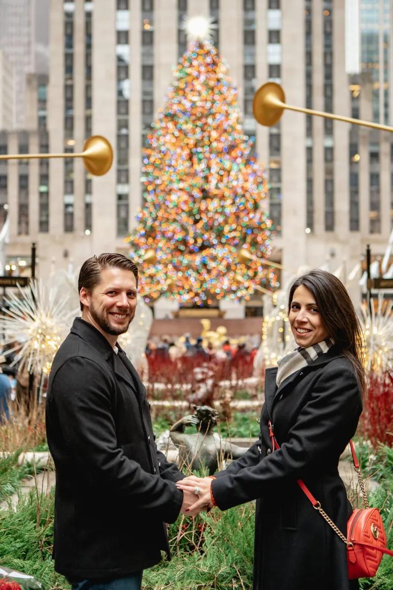 A couple is standing together outdoors in New York during the holiday season. They are holding hands and smiling at the camera. Behind them there is a large Christmas tree decorated with colorful lights, along with festive winter decorations and blurred city buildings. The scene looks warm and joyful, capturing a celebratory moment in a busy holiday setting.