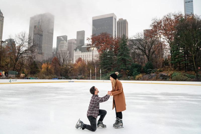 A man proposes to his partner on the ice at Wollman Rink in Central Park. He is kneeling on one knee in ice skates while she smiles and holds his hands. Behind them the New York City skyline and The Plaza Hotel rise above the trees, creating a romantic winter scene in one of the park’s most iconic locations.
