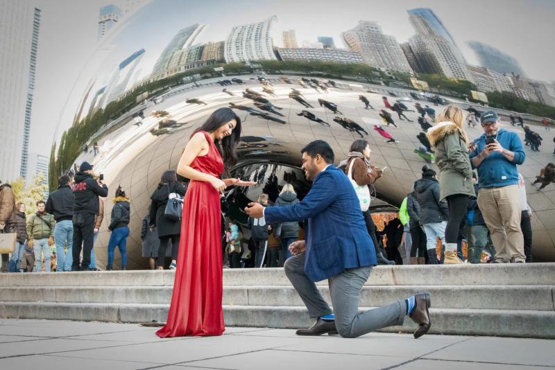 Marriage proposal at The Bean in Chicago’s Millennium Park, man on one knee proposing to woman in red dress with city reflection in background.