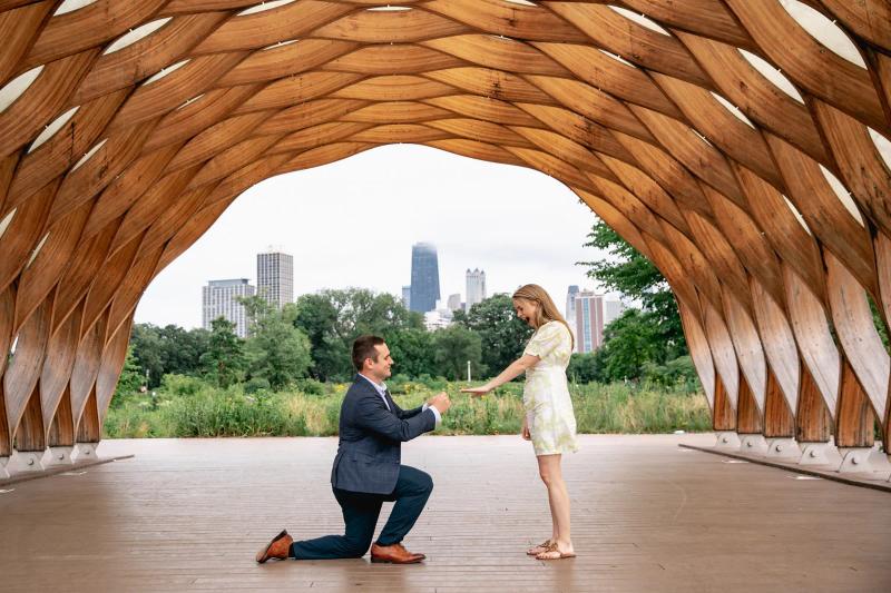 A man proposes to a woman under the Honeycomb Pavilion at Lincoln Park in Chicago, with the city skyline visible in the background.