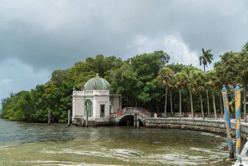Historic waterfront pavilion with a domed roof at Vizcaya Gardens in Miami, surrounded by palm trees and lush greenery, connected by a small arched bridge over calm bay waters under a cloudy sky.