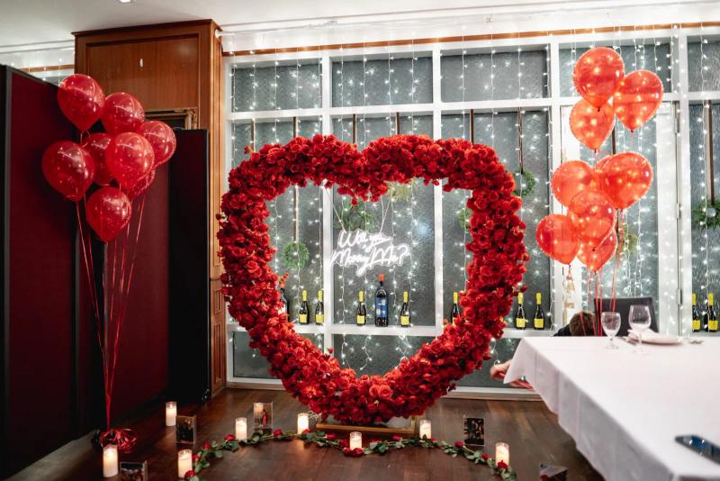 A romantic indoor proposal setup featuring a large heart shaped arrangement of red roses, surrounded by candles, rose petals, and framed photos. Red heart balloons decorate both sides of the space, and a neon sign that reads Will You Marry Me is displayed against a backdrop of twinkling string lights. The room is styled to create an intimate and elegant atmosphere for a private proposal celebration.