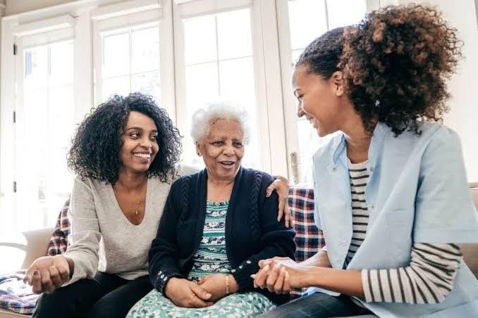 An older woman sits between two younger women on a couch, smiling and talking as they hold hands and one rests an arm around her shoulders.