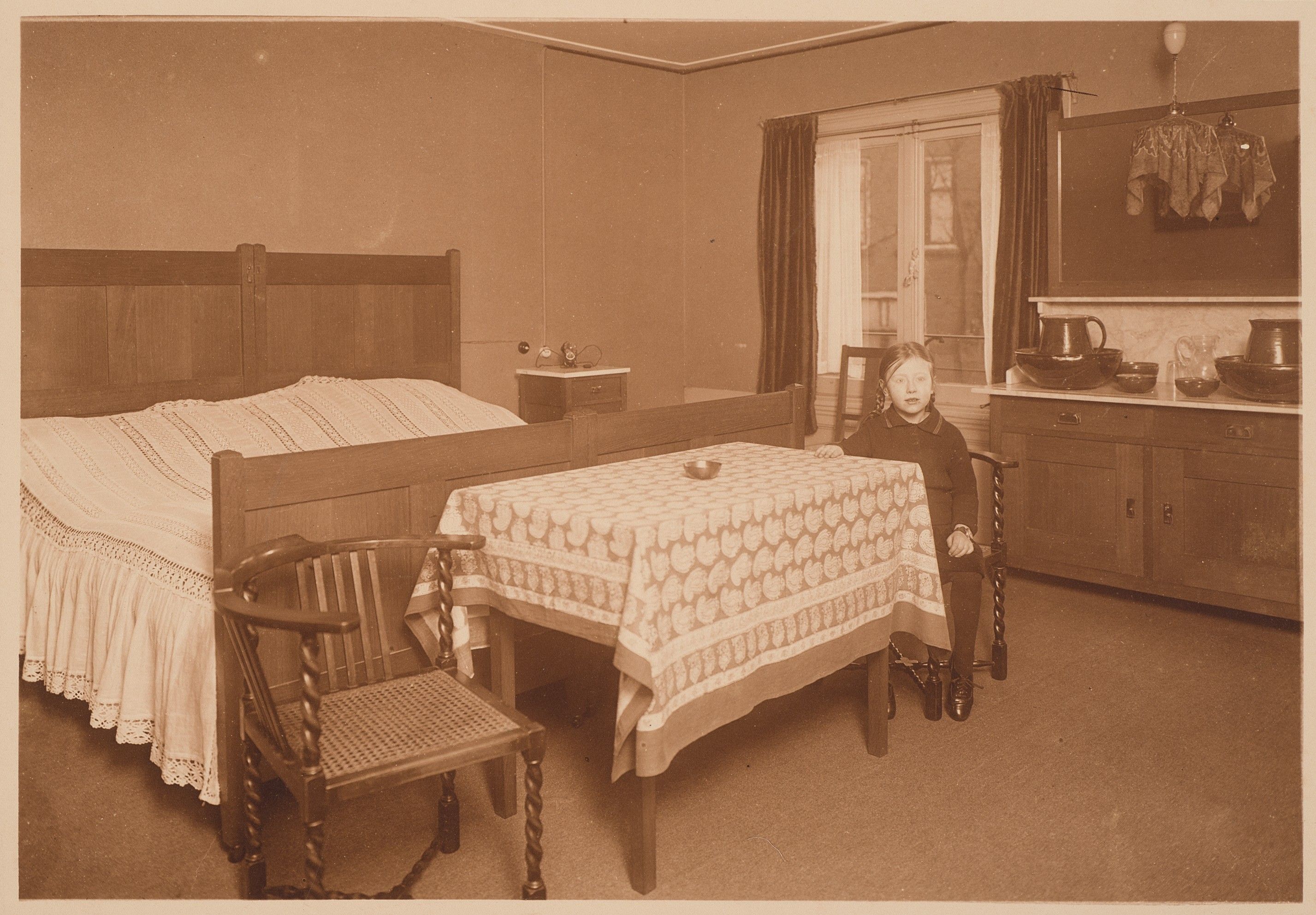 Een meisje, Hanneke Schröder zit aan een tafel met een tafelkleed, in een kamer met een groot houten bed en lage houten kasten.