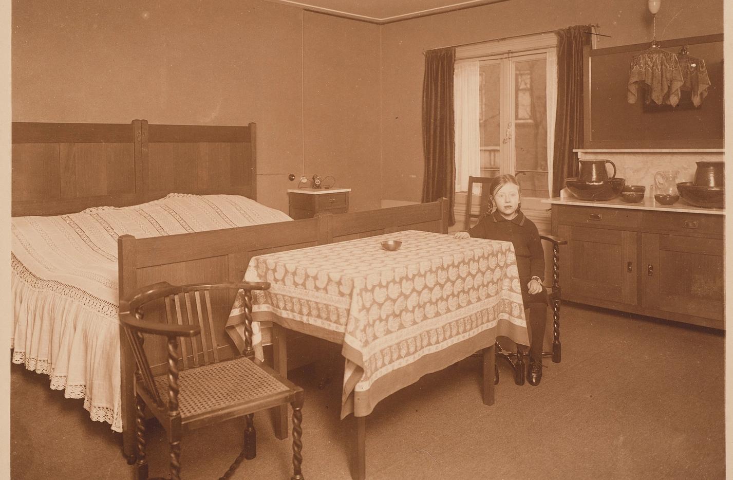 Een meisje, Hanneke Schröder zit aan een tafel met een tafelkleed, in een kamer met een groot houten bed en lage houten kasten.