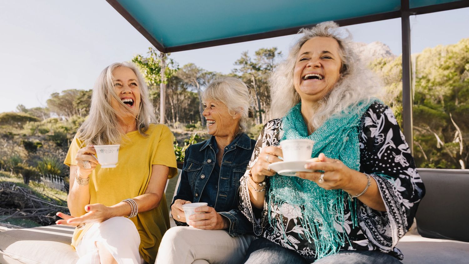 Tre smilende, voksne damer i solskinn, som nyter en kaffekopp sammen i naturen