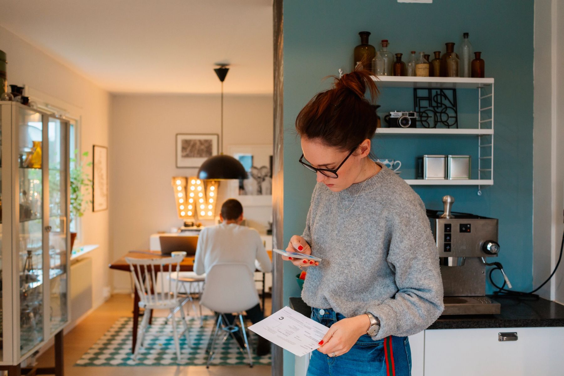 Kvinne står på kjøkkenet og sjekker strømregningen på mobilen, mens en mann jobber ved spisebordet i bakgrunnen. Moderne hjem med hyggelig belysning og kaffemaskin på benken.