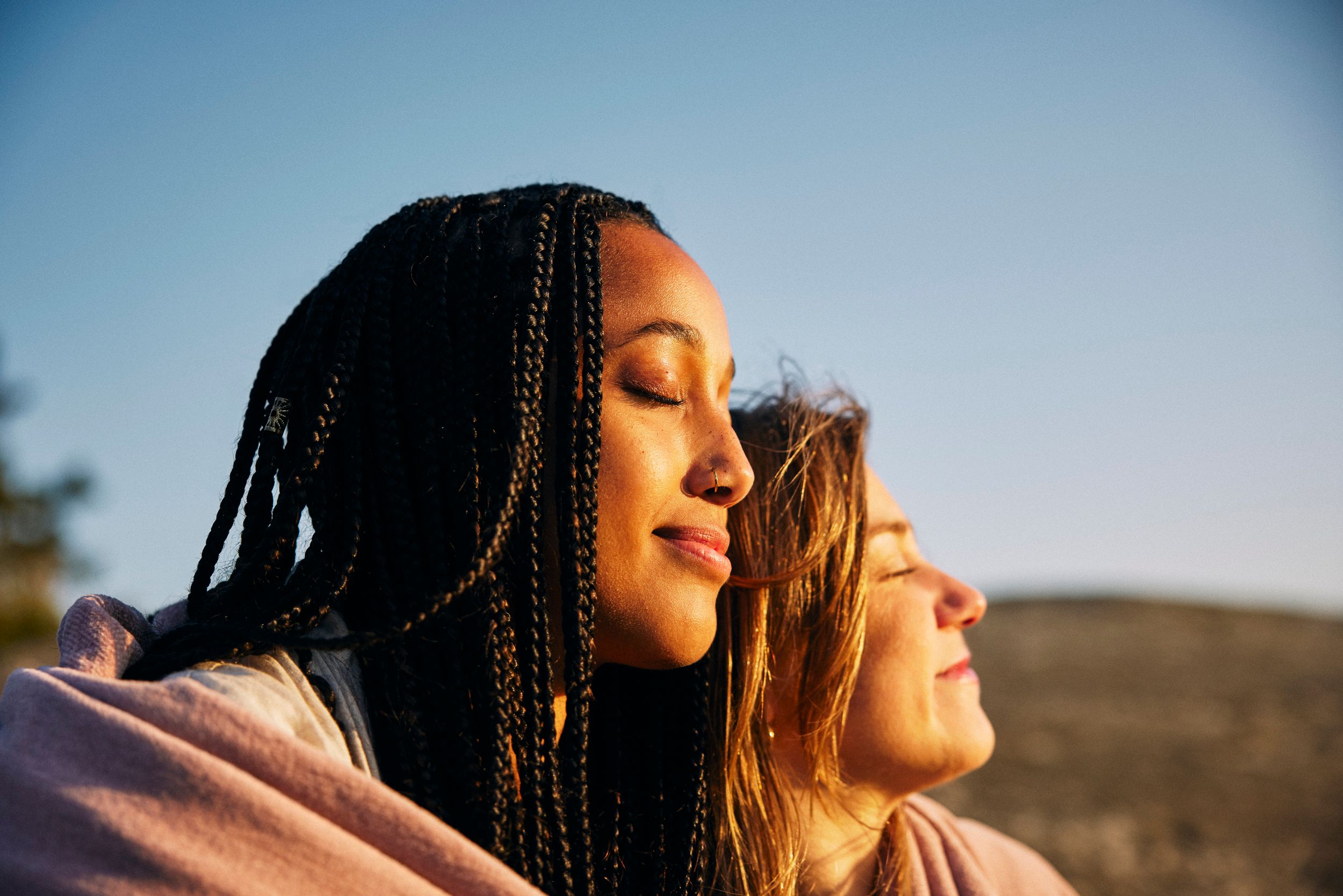 To kvinner nyter solnedgangen sammen, en varm sol skinner i deres ansikter og de lukker øynene mens de nyter solnedgangens magi