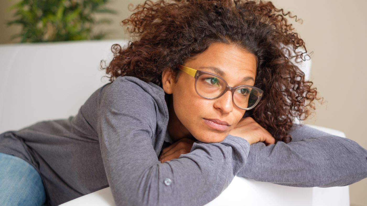 Bekymret, ung kvinne sitter i sofaen. Hun ser oppgitt ut vinduet, og har på seg ledige klær og briller. Hun har en dårlig strømavtale, men skal bytte til Ishavskraft for å få en tryggere, og mer langsiktig strømavtale.