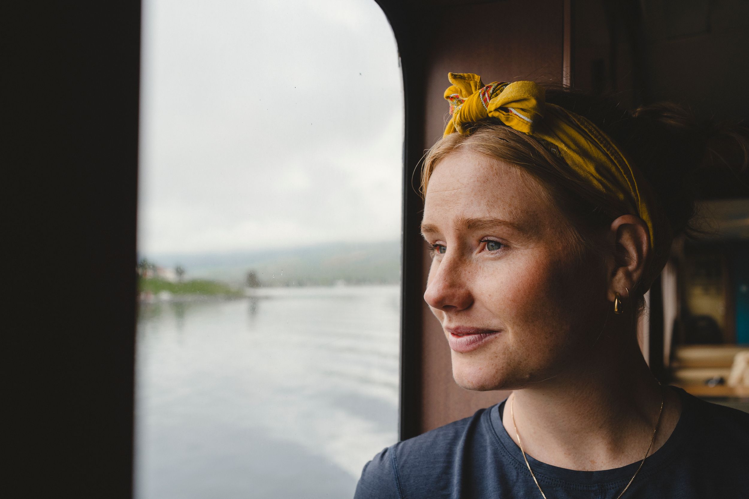 Ei dame med blondt hår ser ut av et vindu på ei ferge, med utsikt over en fjord. Hun smiler beskjedent og har et gult hårbånd på hodet.