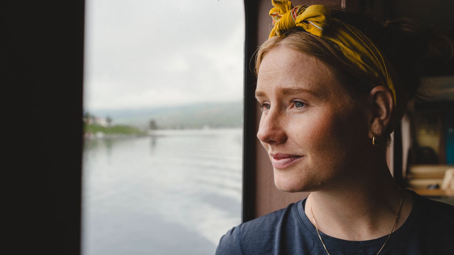 Ei dame med blondt hår ser ut av et vindu på ei ferge, med utsikt over en fjord. Hun smiler beskjedent og har et gult hårbånd på hodet.