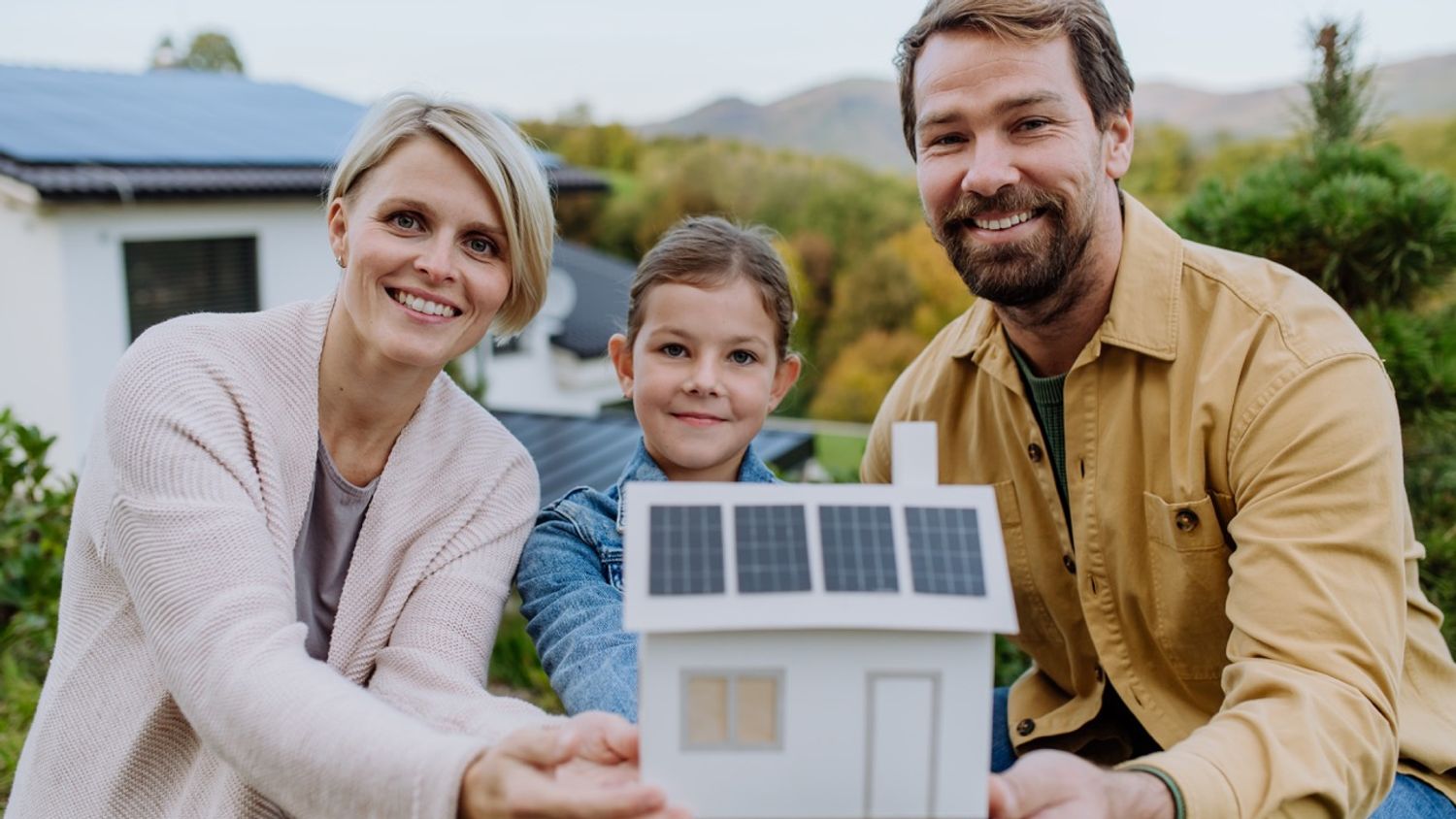 Smilende familie foran huset sitt med solcellepaneler på taket. De holder en miniatyrmodell av et hus med solceller, som symboliserer produksjon av egen strøm.