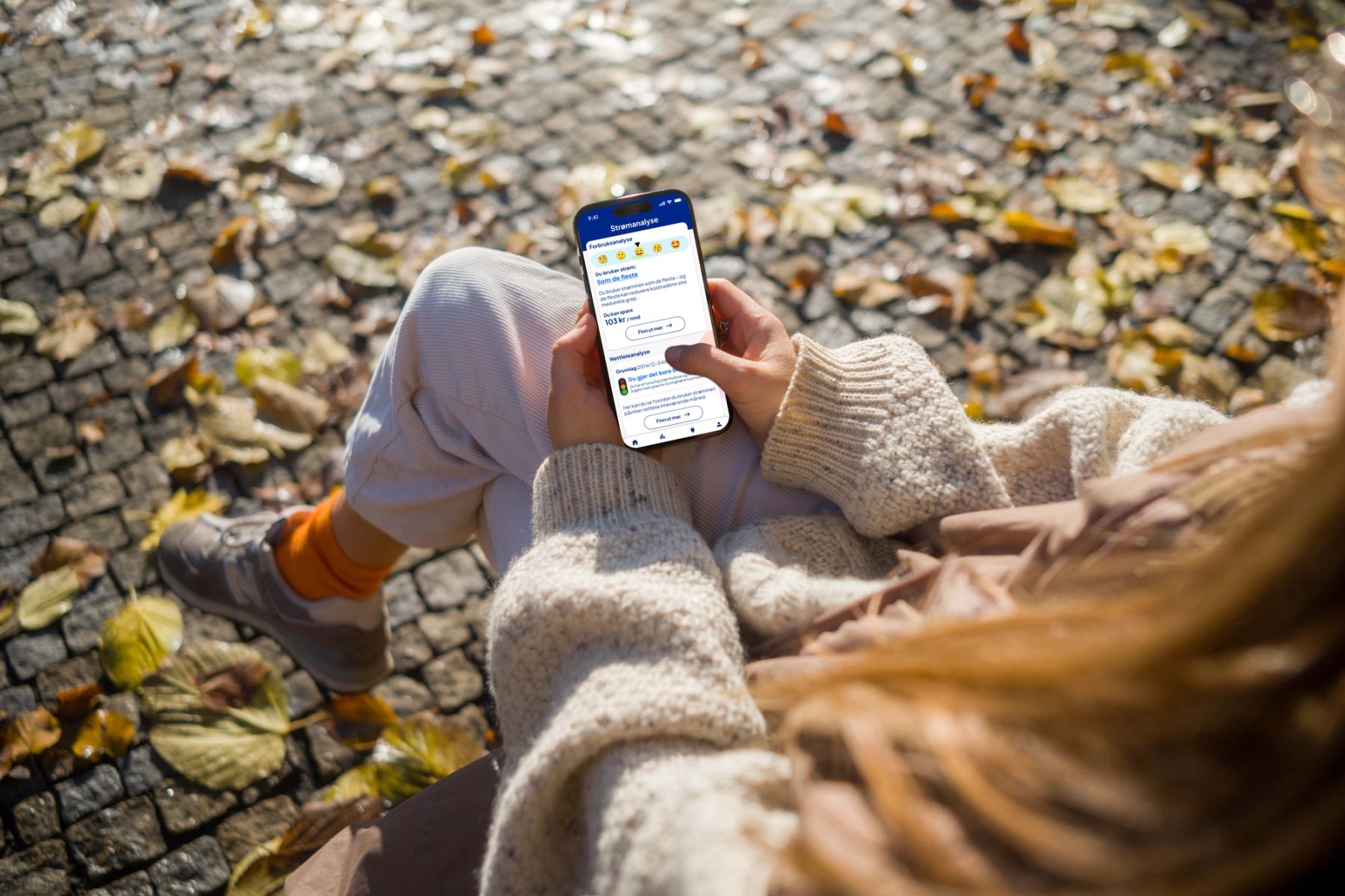 Ung kvinne som sitter ute på en benk i ullgenser på høsten. Det er solskinn og løv på bakken. Hun holder en telefon hvor hun besøker Ishavskraft-appen, og ser på Din strømanalyse.