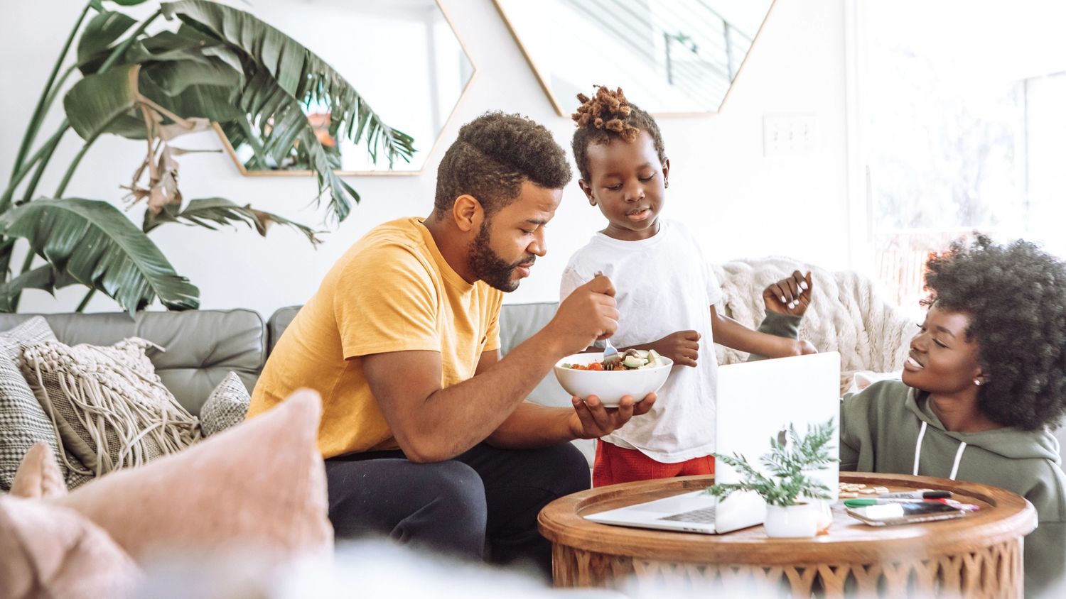 En smilende familie i en lys stue, der far deler snacks fra en bolle mens mor og barn ser på