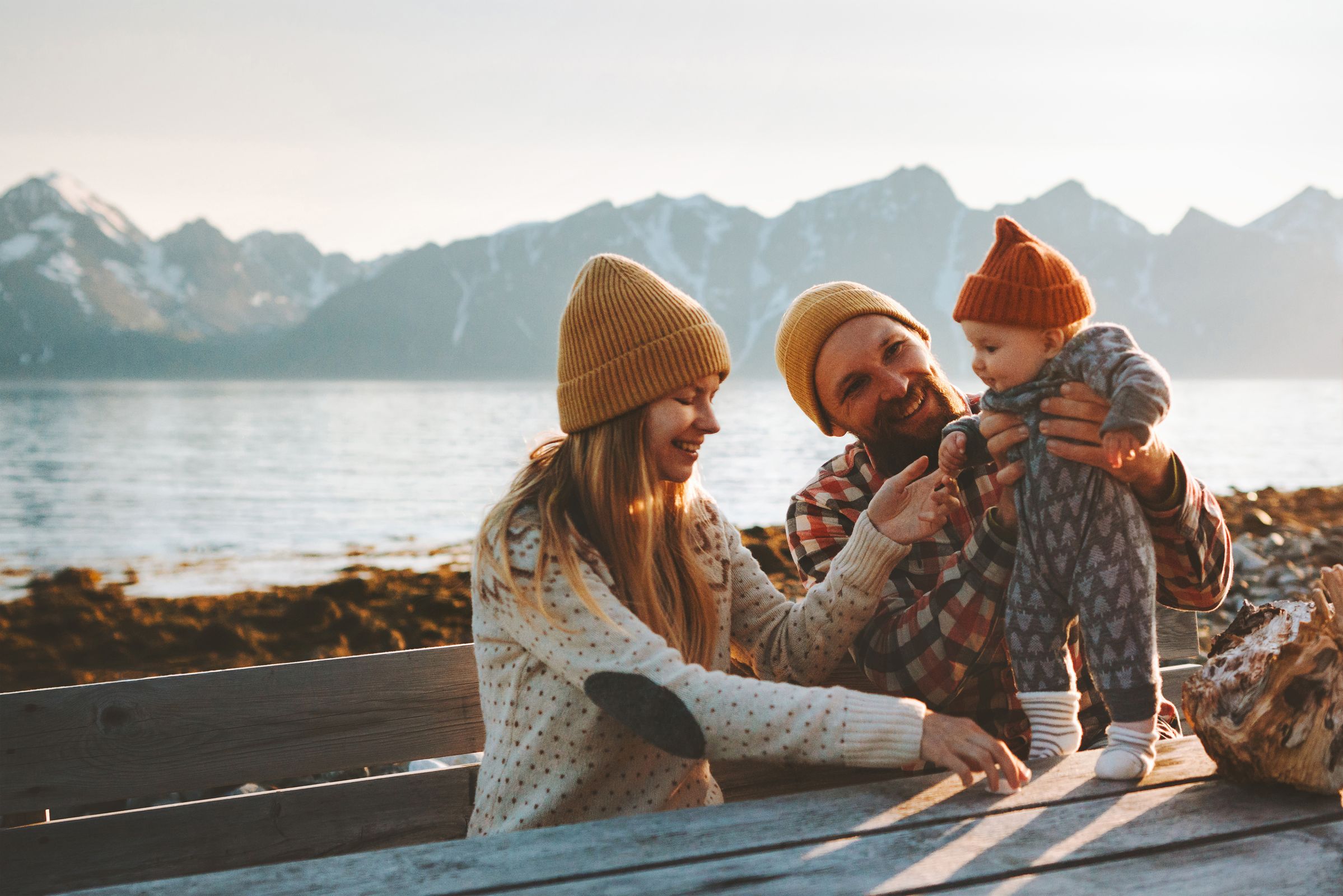 Et ungt par, smilende med sitt lille barn, sitter på en trebenk i fjæra i Nord-Norge med havet og flotte fjell bak seg. De er trygge på Ishavskraft.