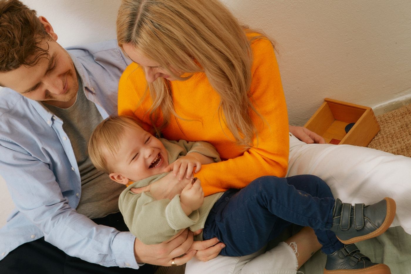 En ung familie leker sammen. Moren ser ned på barnet sitt på 2 år mens hun kiler han, og faren passer på og holder rundt dem. Et flott øyeblikk.