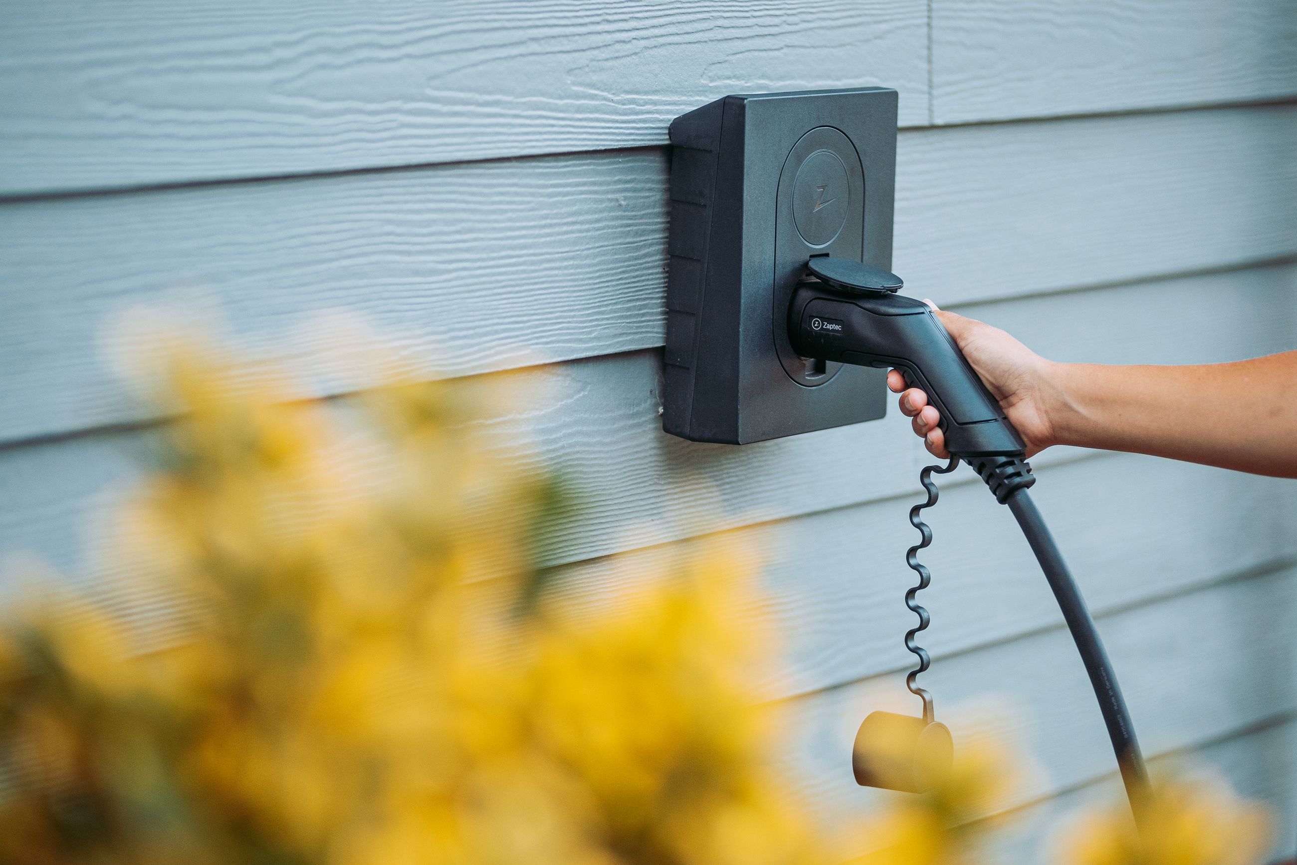 Hånd som kobler til en Zaptec Go elbillader montert på husvegg – trygg og moderne hjemmelading.