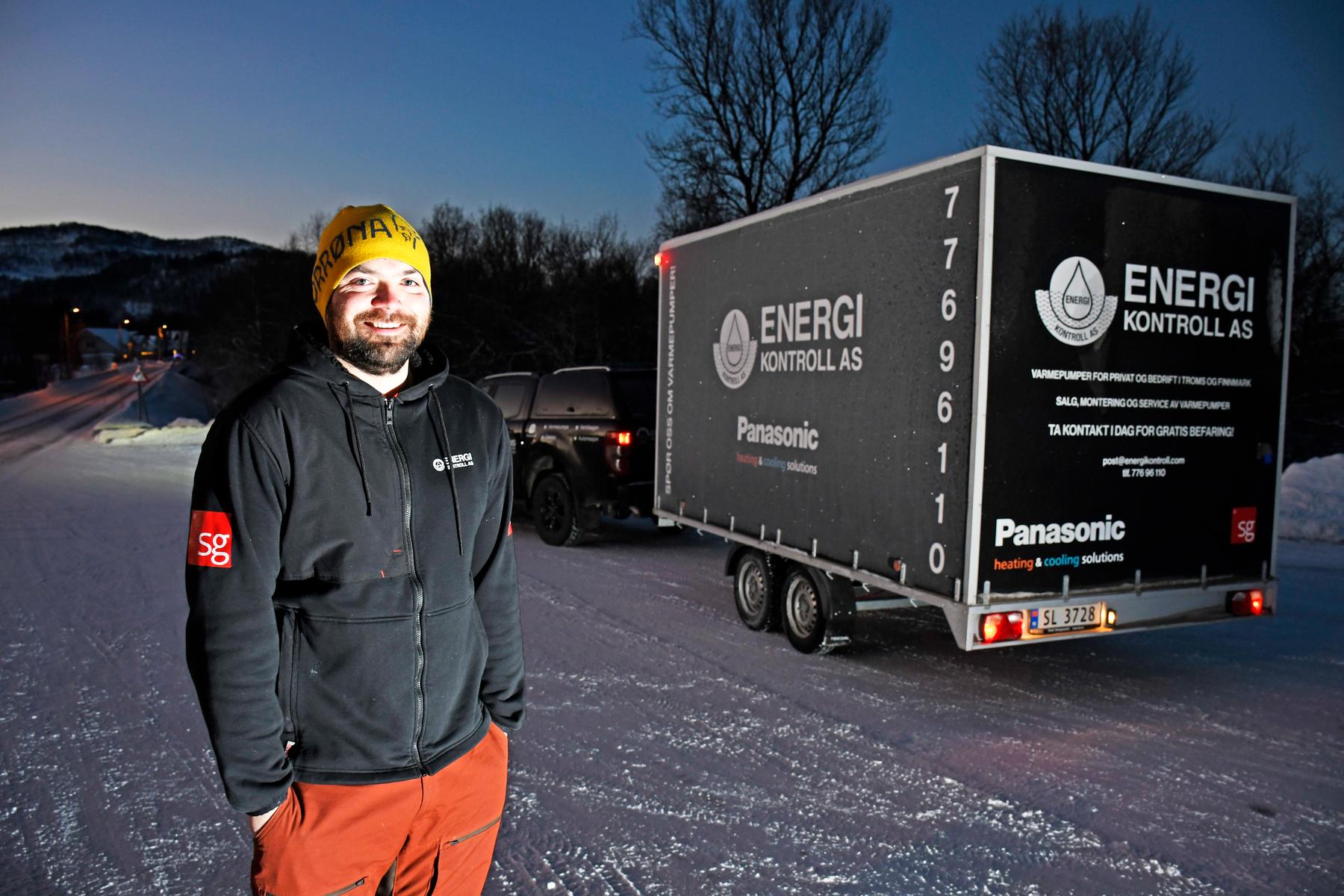Pål-Oskar Guttormsen har i en årrekke montert varmepumper i Troms og Finnmark, her står han smilende ute en tidlig desemberdag foran tilhengeren sin som han bruker på å frakte varmepumpene.