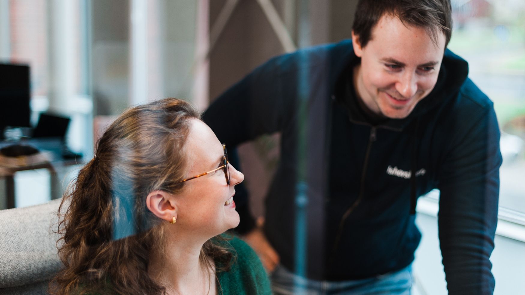 To medarbeidere hos Ishavskraft smiler og ler muntert. Den ene kvinnelige ansatte sitter ved kontorpulten sin, mens den mannlige ansatte står ved siden av og ser mot en laptop-skjerm. Det er lyst og luftig rundt dem, og bildet er tatt foran en glassvegg.