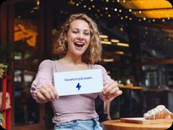 Smilende og glad kvinne som holder opp et gavekort på strøm. Hun har blondt hår til skuldrene, rosa topp og jeans og står i et lokale med julelys og varme farger. En croissant ligger på et bord i forgrunnen.