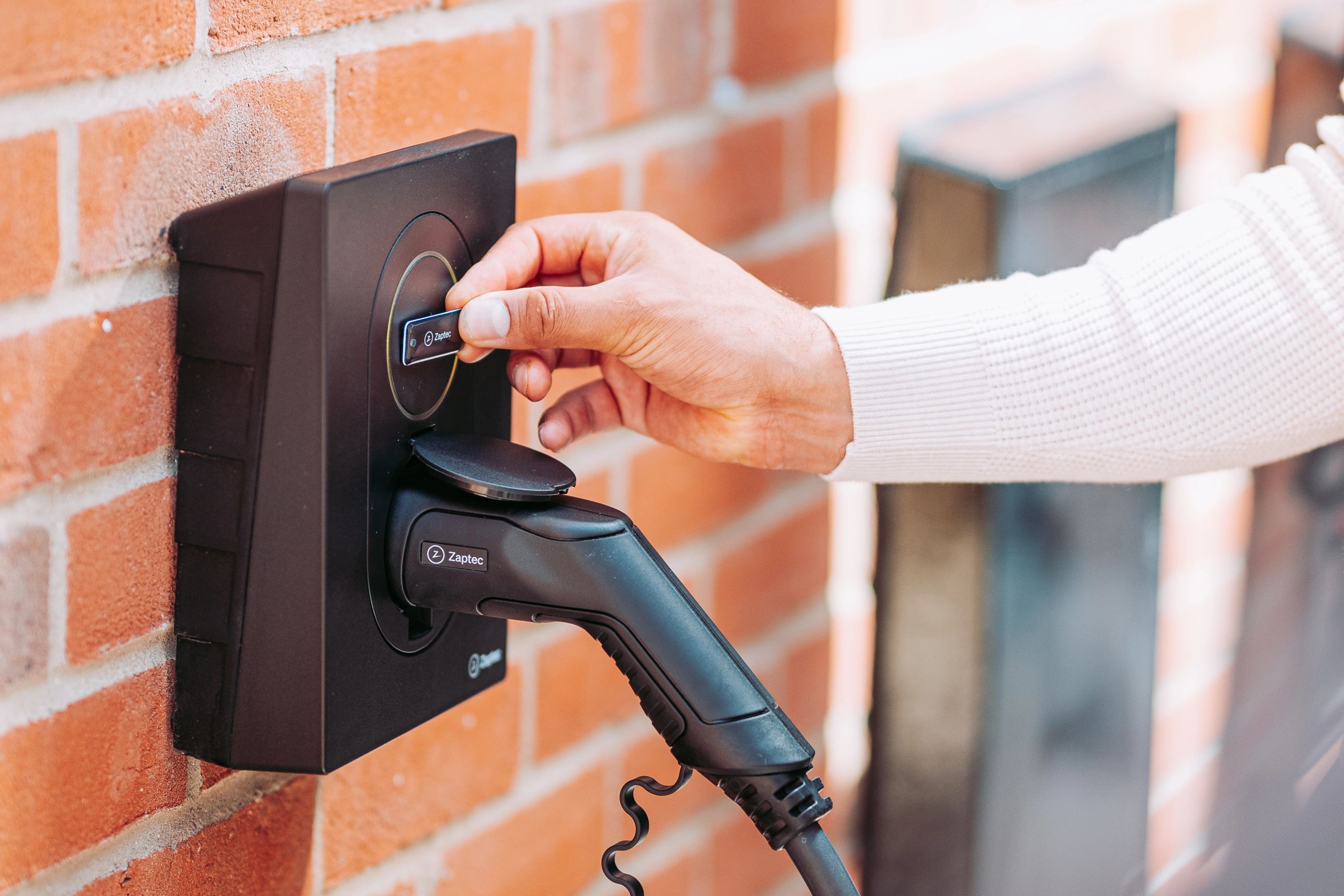 Nærbilde av Zaptec Go elbillader på murvegg – hånd holder Zaptec RFID-brikke for å starte trygg og kontrollert elbillading.