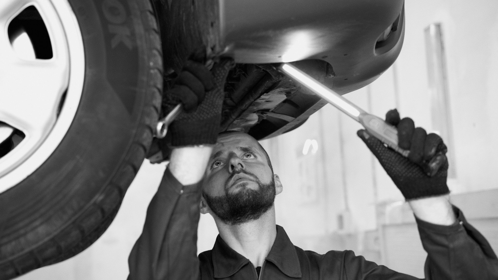A black and white photo of a mechanic inspecting the underside of a car, which is hoisted up.