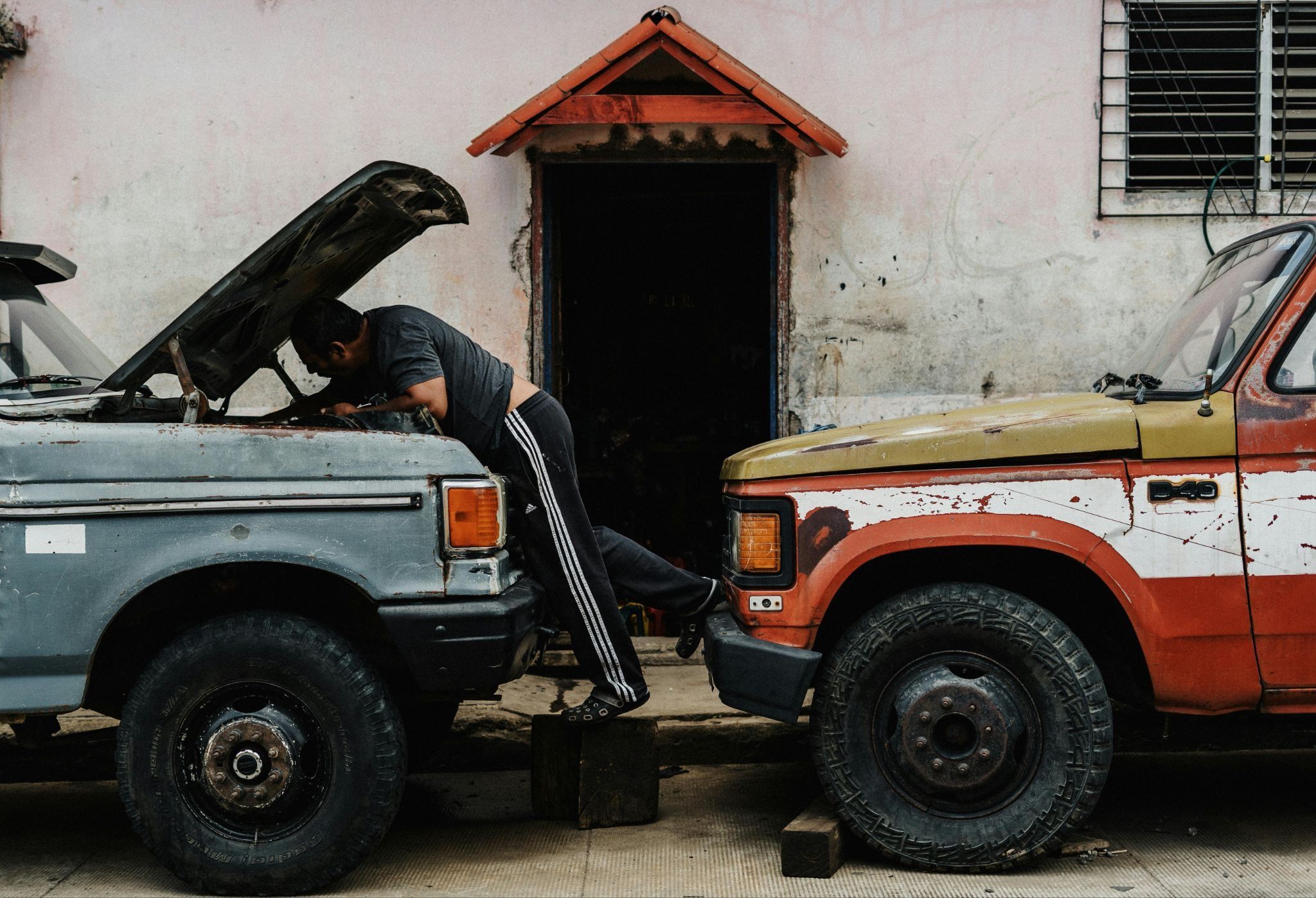 An old red truck and blue truck are parked head to head, with a person fixing something under the hood of the blue truck.
