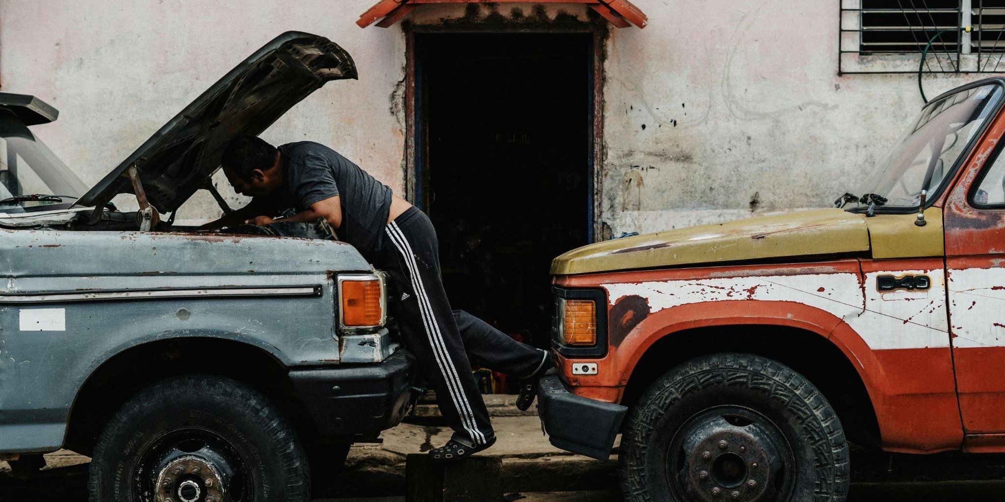 An old red truck and blue truck are parked head to head, with a person fixing something under the hood of the blue truck.