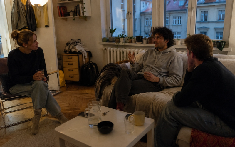 Three roommates relaxing and talking together in a cosy, lamplit shared living room with water glasses on the coffee table