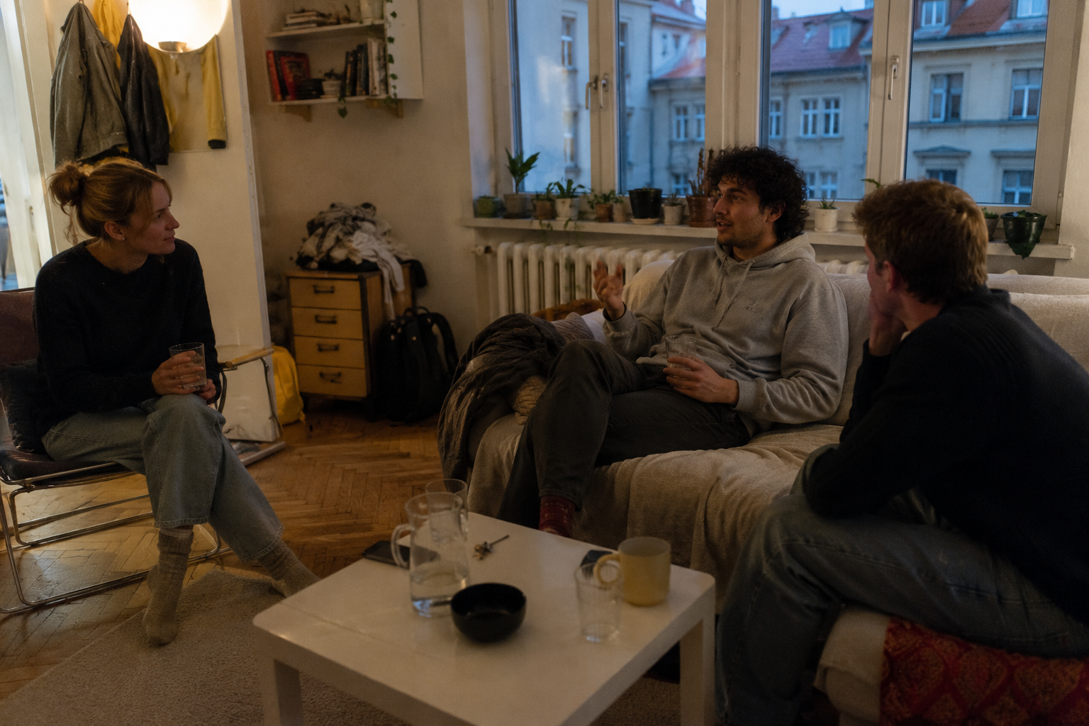 Three roommates relaxing and talking together in a cosy, lamplit shared living room with water glasses on the coffee table