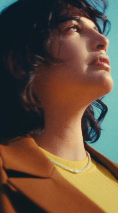 A brunette model wears a lab-grown diamond tennis necklace with a central emerald stone. She is wearing a yellow knit t-shirt with a brown blazer, against a bright blue sky.