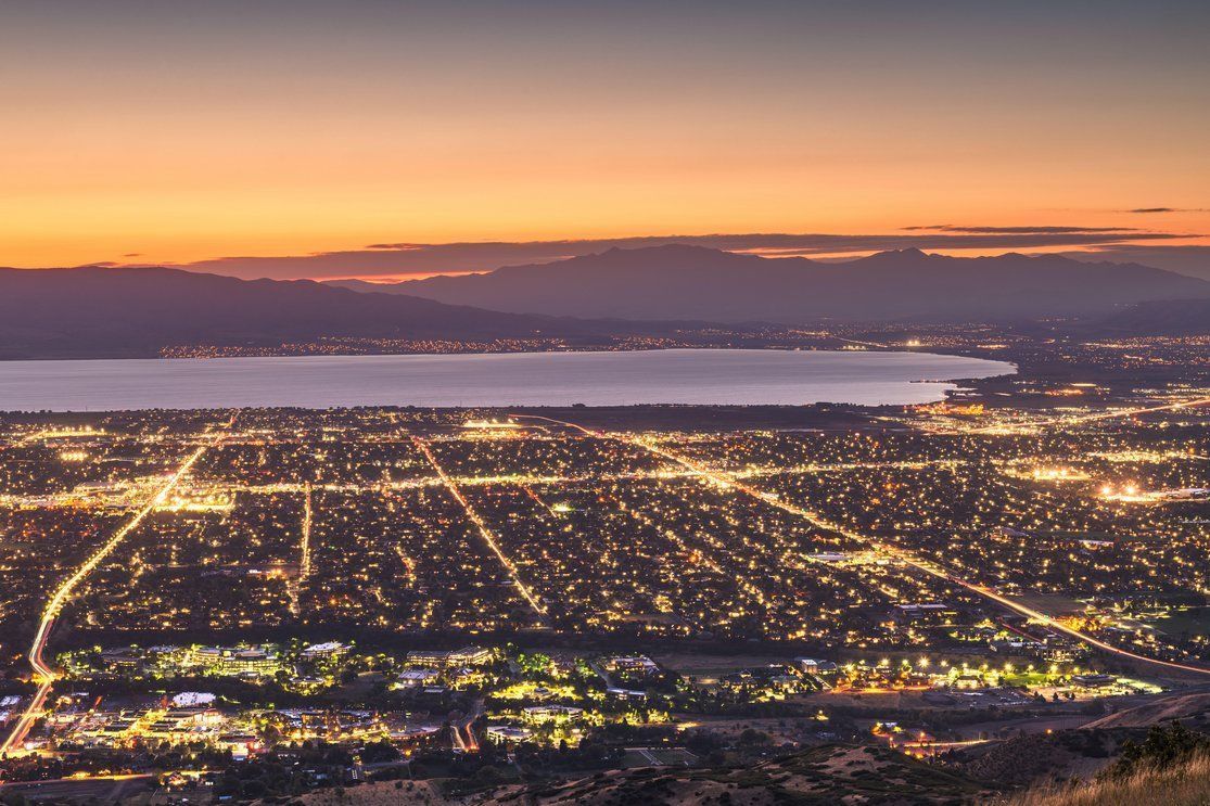 Vue d’une ville illuminée la nuit avec des réverbères et des bâtiments éclairés, surplombant un plan d’eau calme et des montagnes lointaines sous un ciel coloré au coucher du soleil.