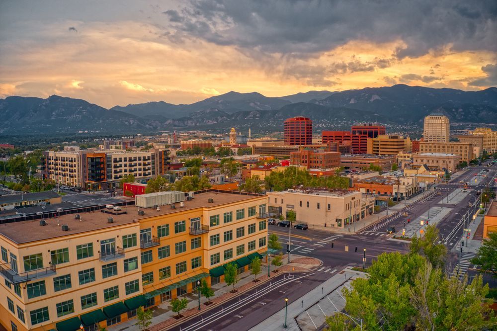 Vue panoramique d’une ville avec des bâtiments modernes et de larges avenues, sur fond de montagnes. La scène est baignée d’une lumière chaude au coucher du soleil, mettant en valeur le paysage urbain.