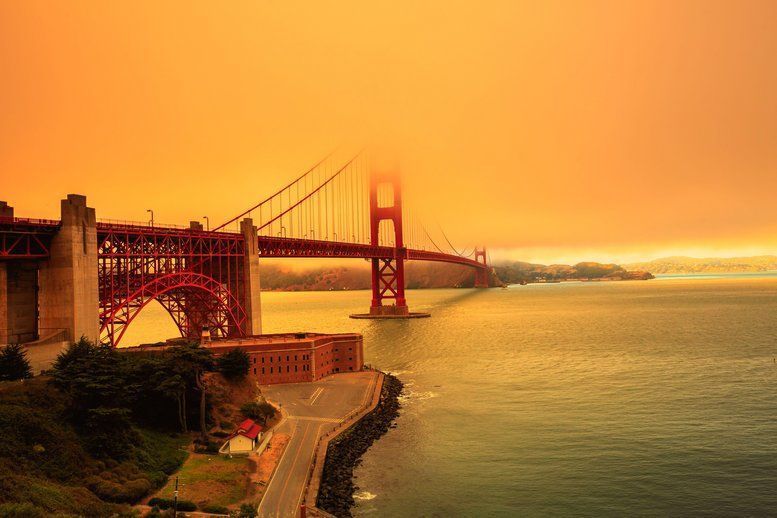 Vue du Golden Gate Bridge partiellement enveloppé de fumée, avec un ciel teinté d’orange et des eaux calmes en dessous, possiblement en raison des incendies à proximité affectant l’atmosphère.