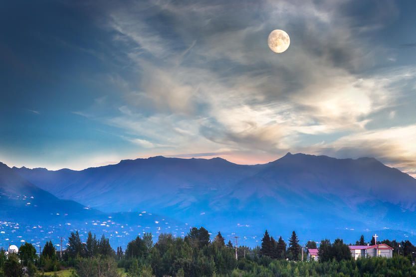 Vue d’un paysage montagneux sous un ciel dégagé avec une pleine lune au‑dessus, diffusant une douce lumière sur la scène. Les montagnes se découpent en silhouette contre le ciel du crépuscule, avec des nuages qui dérivent doucement.