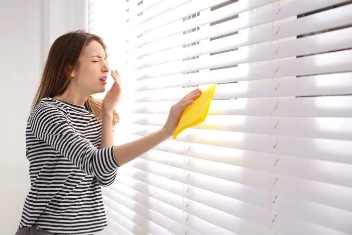 Woman sneezing while dusting window blinds with a yellow cloth.