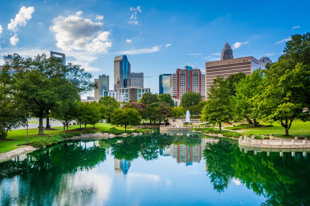 Charlotte skyline reflected in a tranquil pond, surrounded by green trees and blue sky.