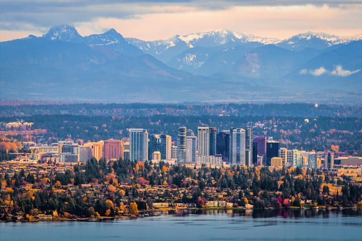 Vue d’un horizon urbain avec de grands immeubles, entouré d’arbres aux couleurs d’automne, sur fond de montagnes enneigées et d’un lac calme.