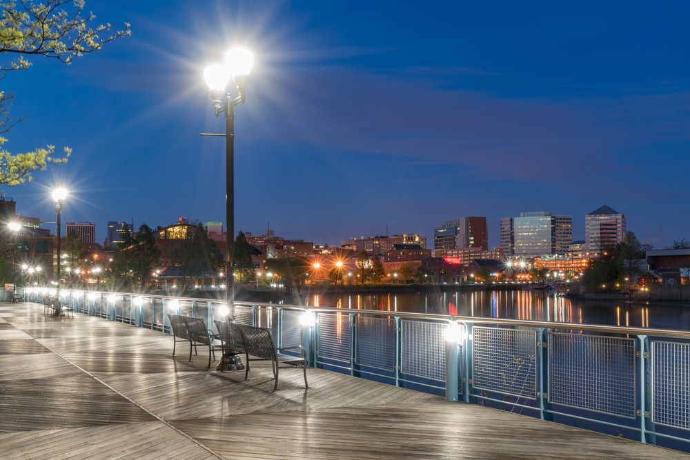 Wooden boardwalk lined with benches and lampposts overlooking a glowing city skyline across the water at dusk.