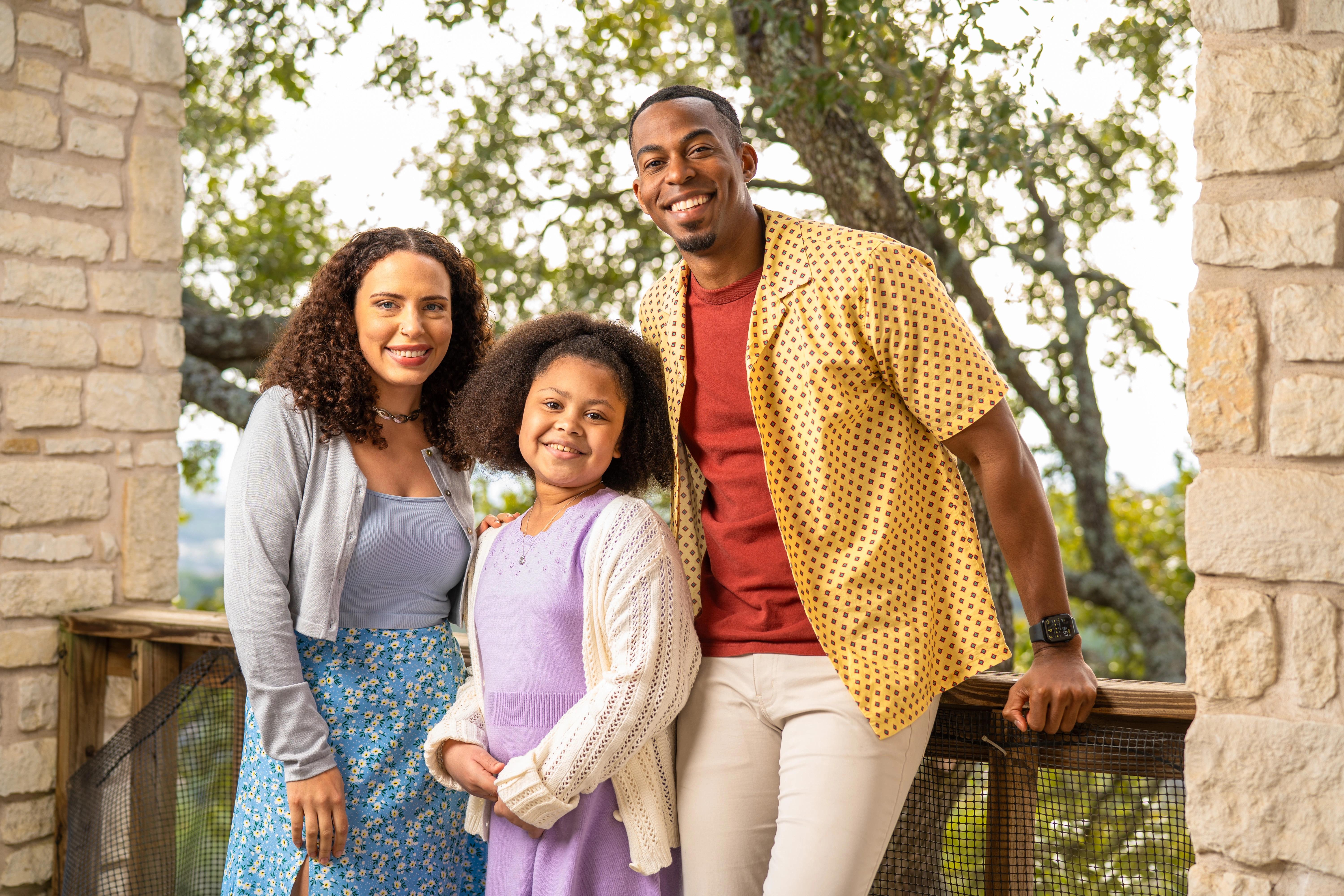 A smiling family of three standing together on a rustic wooden patio, surrounded by greenery and stone columns.