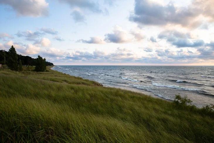View of a grassy shoreline along a large lake with gentle waves and a partly cloudy sky at sunset.