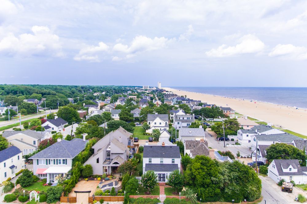 Aerial view of a coastal town with residential homes and a sandy beach stretching along the shoreline under a partly cloudy sky.