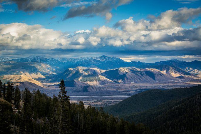 Vue d’une vallée montagneuse avec des versants boisés au premier plan et des sommets lointains sous un ciel partiellement nuageux et dramatique.