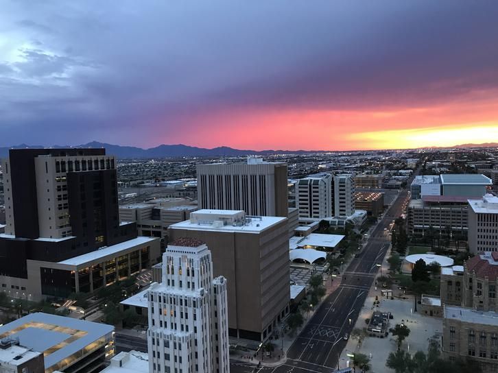 Vue d’une ville au coucher du soleil, avec de grands immeubles et des rues visibles, et un ciel coloré aux teintes vibrantes d’orange, de violet et de rose à l’horizon derrière les montagnes.